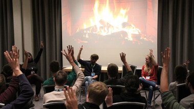 Vortragsszene mit animiertem Lagerfeuer auf Leinwand und erhobenen Händen im Publikum.